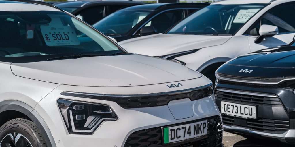 Row of modern electric cars parked on a dealership forecourt, featuring a white Kia with a green EV license plate and a sold sign in the windscreen.