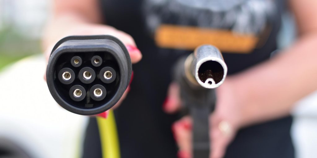 Hand holding a black Type 2 EV charging cable next to a fuel pump nozzle, comparing electric vehicle charging with petrol fueling.